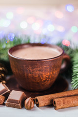 Blurred Christmas lights, cup of hot cocoa with milk, broken chocolate cubes, hazelnut and cinnamon sticks, christmas tree branches, pine cone, selective focus
