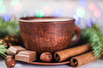 Blurred Christmas lights, cup of hot cocoa with milk, broken chocolate cubes, hazelnut and cinnamon sticks, christmas tree branches, pine cone, selective focus
