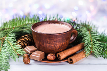 Obraz na płótnie Canvas Blurred Christmas lights, cup of hot cocoa with milk, broken chocolate cubes, hazelnut and cinnamon sticks, christmas tree branches, pine cone on white wooden planks