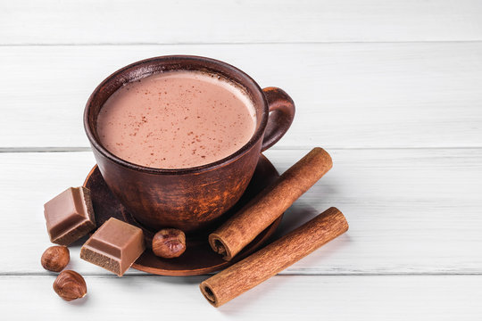 Hot cocoa with milk in brown clay cup, broken chocolate cubes, hazelnut and cinnamon sticks on white wooden planks