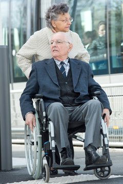 elderly man seated in wheelchair pushed by his wife