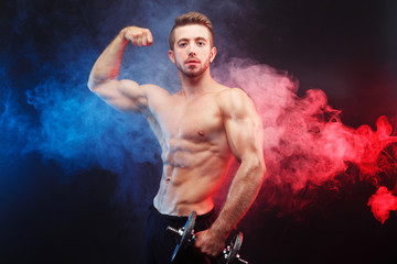 Fototapeta na wymiar Strong athletic young man with huge muscles posing with dumbbell in hand on dark studio background