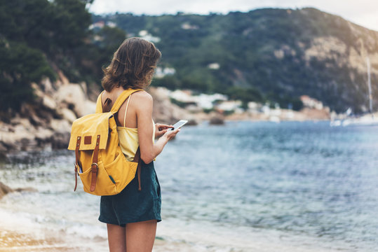 Hipster girl with backpack hold on smart phone gadget in sand coastline. Traveler using in female hand mobile on background beach seascape horizon. Tourist look on blue sun ocean, summer lifestyle