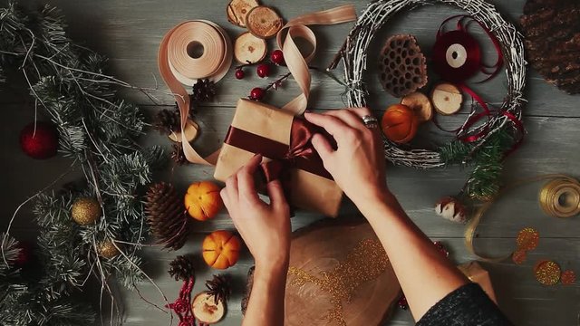 A top-down plan. Fully visible the table with the decorations. Female hands put and finalize Christmas gift wrapped in craftool paper on a wooden table. Bandaging tape and tied in a bow.