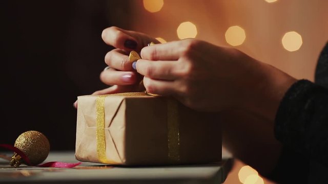 Close-up on the background of Christmas lights women's hands tying the ribbon on the gift of Kraft paper. The background is blurred in bokeh.