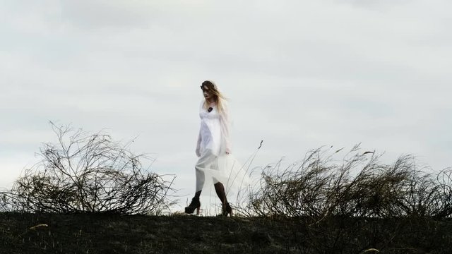The young woman with spooky makeup for Halloween in a white bride dress walking through the field of dried grass on the background of the sky. 4K