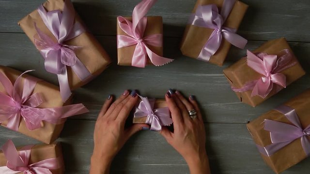 A top-down plan. Fully visible the table with the decorations. Female hands put and finalize Christmas gift wrapped in craftool paper on a wooden table. Bandaging tape and tied in a bow.