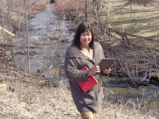 Professional looking mid-aged Filipina standing beside a fast moving drainage ditch.    with note book and recording device in early spring.