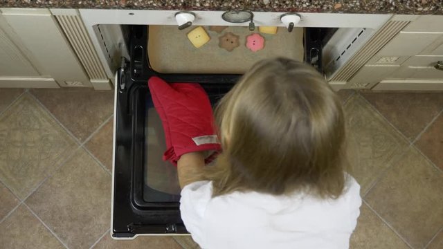 Top view of young white blonde girl pushing tray with ginger cookies bisquits to oven at home kitchen. High angle top view. CU. Red mitten mitt glves.