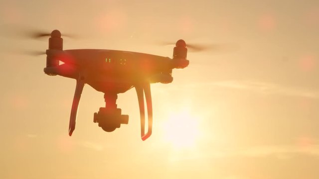 CLOSE UP LENS FLARE SILHOUETTE Small filming drone flying over golden sunset sky. White quadcopter drone with attached camera filming at golden light. Quadrocopter helicopter flying across sunrise sky
