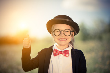 Outdoor portrait of funny little girl in bow tie and bowler hat showing thumb up. Retro stile.