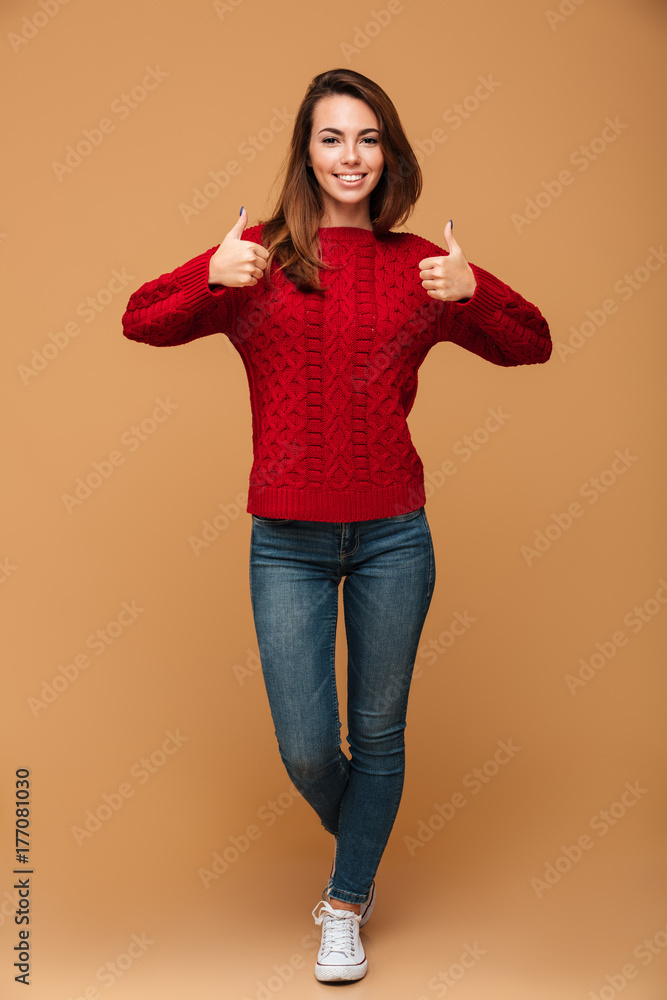 Sticker full length portrait of smiling brunette woman in red knitted sweater and jeans showing thumbs up ge