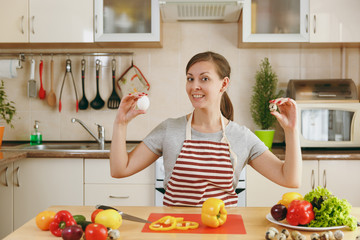 A young attractive pensive woman in an apron chooses between chicken and quail eggs in the kitchen. Dieting concept. Healthy lifestyle. Cooking at home. Prepare food.