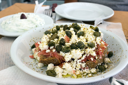 Dakos greek appetizer, dried bread, tomatoes, feta, or mizithra cheese, capers, oregano; Tzatziki in background. Taken in Naxos