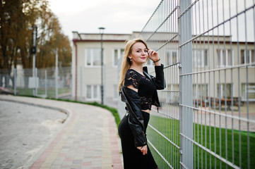 Elegant blonde girl wear on black posing at streets of town against iron cage.