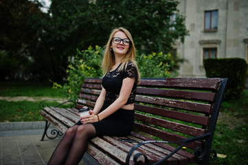 Elegant blonde girl wear on black posing at streets of town, sitting on bench with cup of coffee at hands.