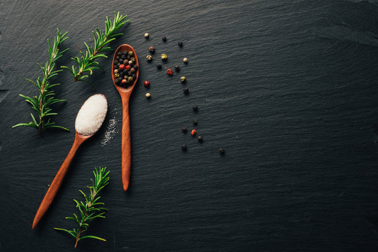 Mix of colorful spices in wooden spoons. Sea salt, mixed pepper, and fresh rosemary on black slate background. Top view. Copy space.