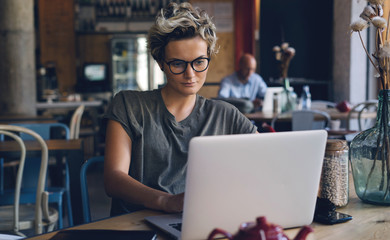 Young attractive caucasian female wearing glasses is typing messages on a laptop connected to...