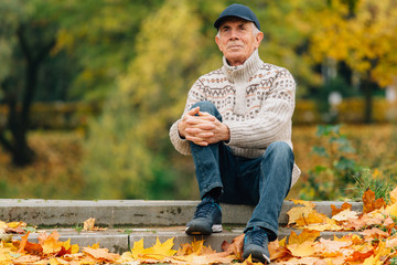 Portrait of pretty senior man in cap sitting in colorful autumn park. Pensioner resting outdoors....