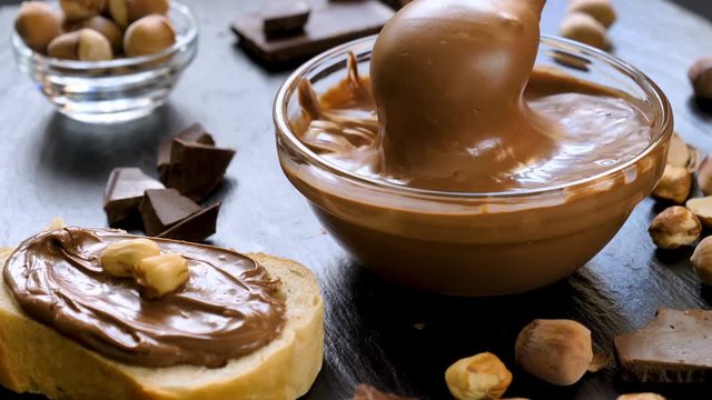 Chocolate spread in glass bowl with hazelnuts and cream on slice of bread on black table. Serving a delicious breakfast concept.