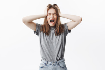 Young beautiful european student with dark hair in grey t-shirt and jeans holding head with hands, screaming with closed eyes,after being attacked with big amount of work