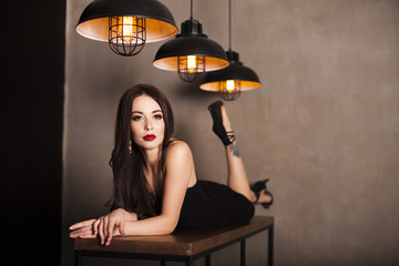 Young beautiful sexy brunette woman wearing black evening dress with decollete, makeup with red lips and hairstyling lying on a table. Fashion vogue style portrait.