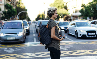 Attractive girl with short blonde hair wearing casual clothes with a backpack on her back, blank paper cup and mobile phone in her hand is crossing the road on a blurred urban traffic background.