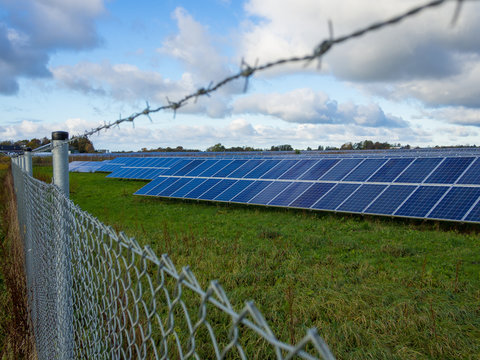 Solar Panel Or Photovoltaic Farm Behind Metal Chainlink Fence On Green Field With Dramatic Cloudy Sky In North Germany