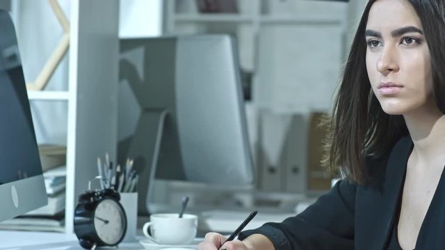 Cinemagraph of young pretty businesswoman with hair flowing in the air sitting at desk in the office, holding pen and looking at computer screen