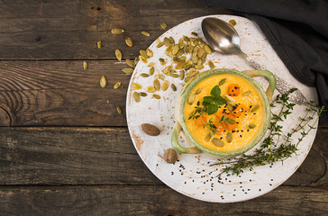 Pumpkin soup in beige bowl with pieces of pumpkin, seeds and herbs on old wooden background.