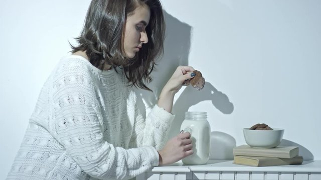 Cinemagraph of young brunette woman in white woolen sweater sitting at kitchen table and holding dunked cookie above glass jar with milk