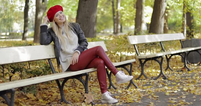 Attractive woman in black leather jacket and red beret sitting on bench in autumn park, leaning her head on hand and smiling.