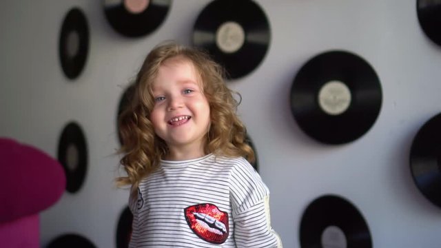The little lovely girl faces the camera in studio. The fair-haired girl costs on a blue background. The beautiful little girl with ringlets smiles in front of the camera. Young kid sticking out tongue