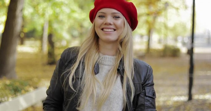 Attractive young woman in red beret and leather jacket standing on alley and looking at camera during her walk in autumn park.