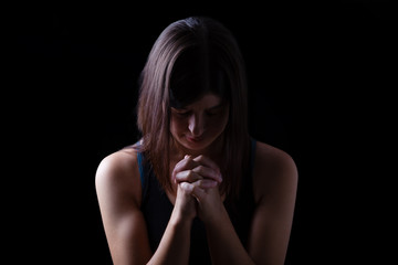 Faithful athletic woman praying, with hands folded in worship to god, head down and eyes closed in religious fervor, on low key black background. Concept for religion, faith, prayer and spirituality
