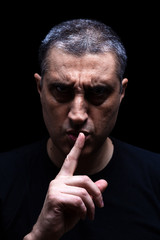 Angry mature man with an aggressive look making the silence sign in a threatening and creepy way. Low key, black background. Concept for secret, threat, anger, rage, violence, danger, menace.