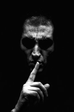 Angry mature man with an aggressive look making the silence sign in a threatening and creepy way. Black and White, black background. Concept for secret, threat, anger, rage, violence, danger, menace.