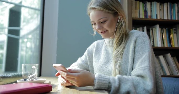 Attractive young woman in soft jumper sitting in front of window, using her smartphone, listening to music and smiling.