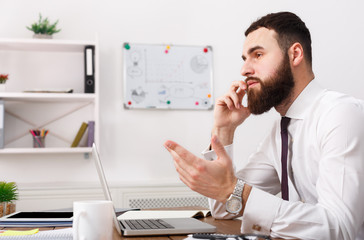 Serious businessman talking on cell phone with laptop in modern office