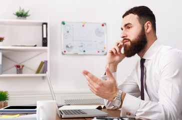 Fototapeta na wymiar Serious businessman talking on cell phone with laptop in modern office