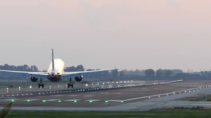 Big plane landing at Barcelona airport seen from behind at sunset with lit signal lights on the runway. 