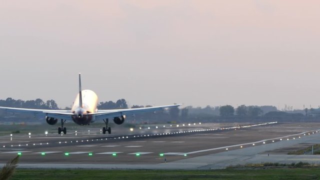 Big plane landing at Barcelona airport seen from behind at sunset with lit signal lights on the runway. 