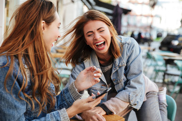 Two overjoyed brunette woman in jeans wear laughing while sitting at city cafe - obrazy, fototapety, plakaty