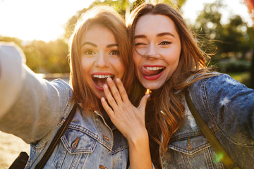 Two cheerful young brunette woman in jeans wear having fun while taking selfie on mobile phone,...