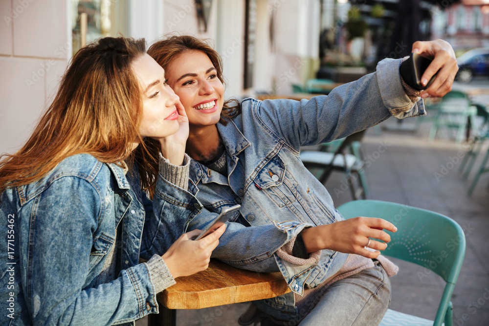 Poster Photo of two charming brunette woman taking selfie on mobile phone while sitting in city cafe