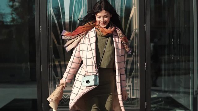 charming young woman walks through the autumn city in a coat and smiles. cute girl with a bouquet of flowers on a background of modern architecture. slow motion