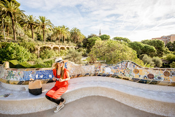 Naklejka premium Young woman tourist in red dress sitting on the bench decorated with mosaic in the famous Guell park in Barcelona. Wide angle image with copy space