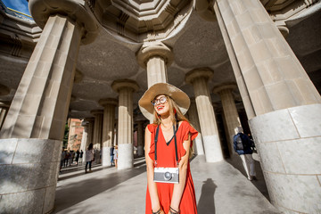 Fototapeta premium Happy woman tourist in red dress with hat standing near the columns visiting famous Guell park in Barcelona