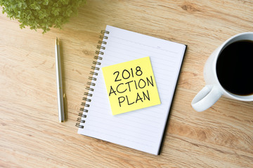 2018 action plan phrase on sticky paper on top of notepad with pen, cup of coffee and flower, wooden table background.