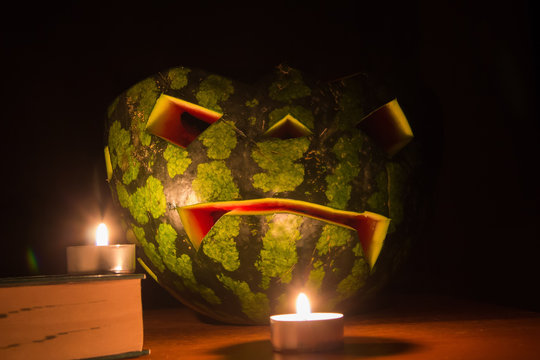 Halloween symbol, water-melon with carved red smiling face. Burning candles and books on wooden table, dark background. Funny, handmade helloween celebrating concept. 
